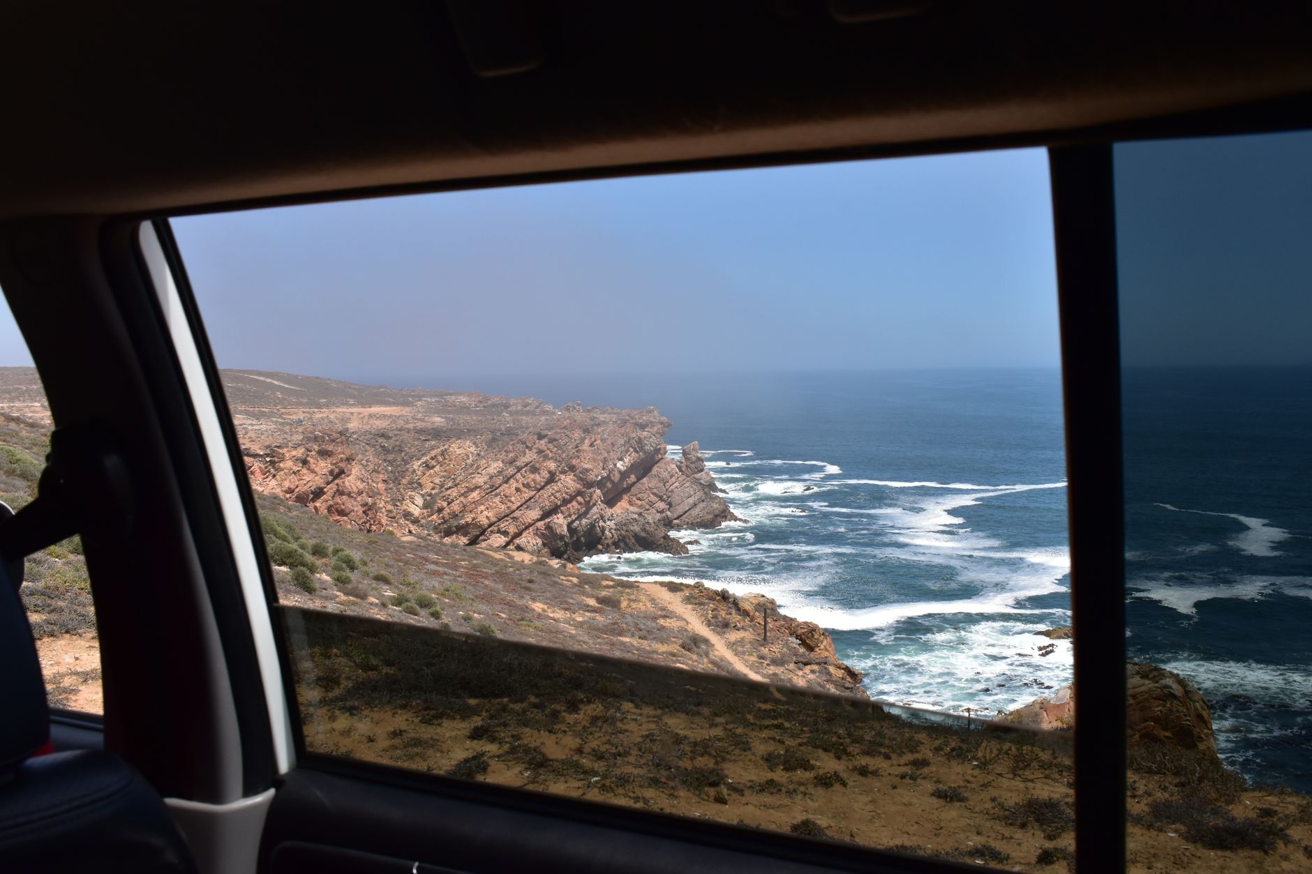 Dramatic coastline seen through a car window, capturing South Africa's rugged cliffs and ocean waves.